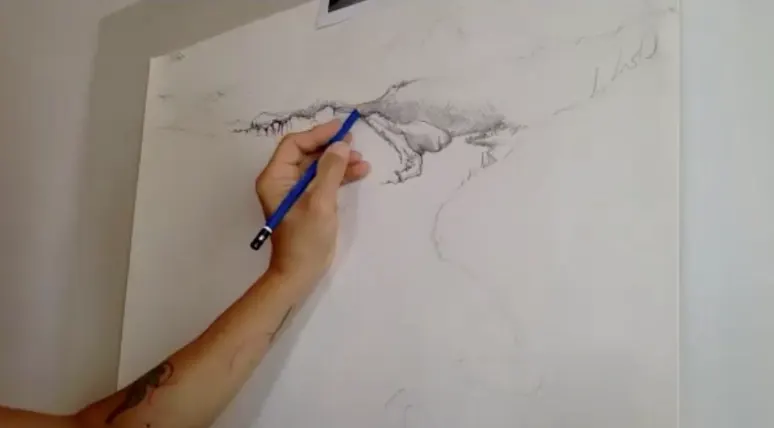 Close-up of an artist sketching a detailed landscape scene with a blue pencil on a large white canvas.